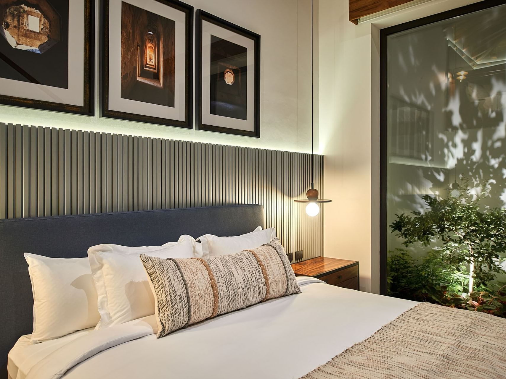 A bedroom with a bed, three framed pictures, a wall light, and a glass wall showing an outdoor garden.