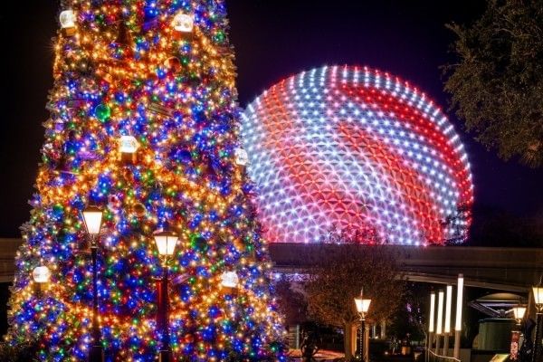 Spaceship Earth at EPCOT lit up as a giant peppermint behind a colorfully lit Christmas tree at night. 