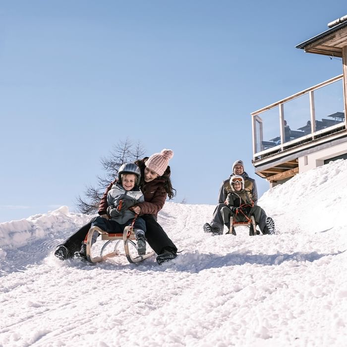 Zwei Familien rodeln einen schneebedeckten Hang hinunter, während andere auf einer Terrasse speisen.