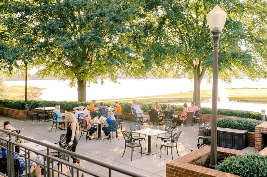 Guests dining at outdoor patio tables beneath shaded trees beside the lake at Clemson University Conference Center & Inn