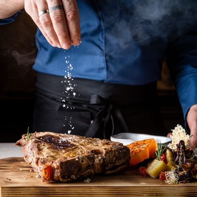 Chef seasoning steak on a wooden board at 12 Hoefe – La cucina dei 12 masi, Falkensteiner Family Resort Lido, Ehrenburg.