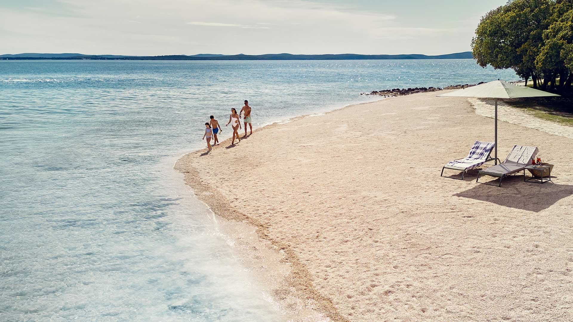 Familie spaziert am Strand, Liegen und Sonnenschirm unter Bäumen, Ozean und Horizont im Hintergrund.
