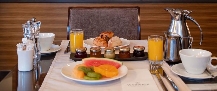 Warwick Geneva breakfast setup with croissants, jam jars, juice, coffee cups, & a fresh fruit plate at Téséo Restaurant & Bar