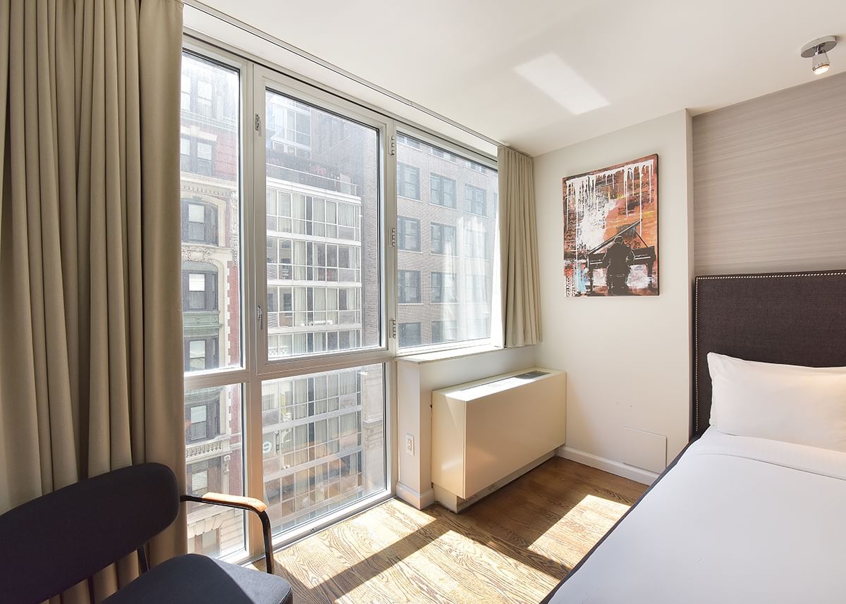 A hotel room with a large window, a bed, a chair, and a painting on the wall.