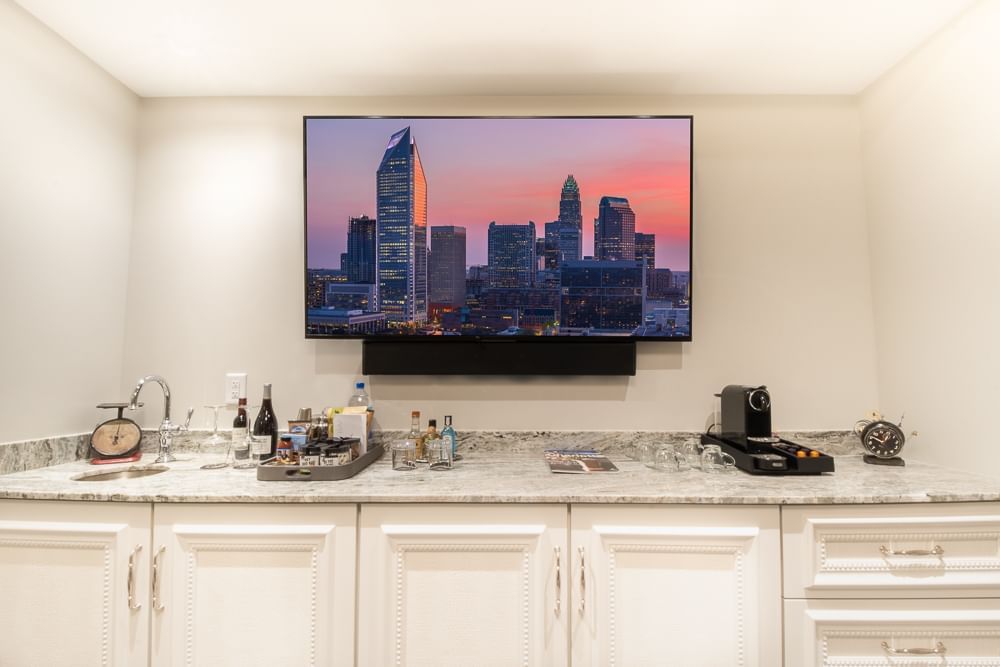 A wide view of a hotel room's wet bar with a large flat screen TV showing a city skyline, white cabinets, and a marble countertop.