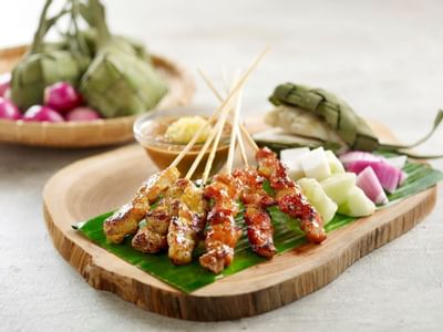 Grilled skewered meat with dipping sauce and fresh garnishes on a wooden platter at York Hotel Singapore