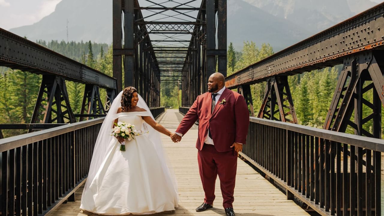 Bride and groom holding hands on a bridge with mountains in the background.