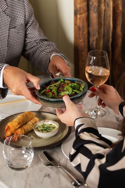 Man serving a salad to a woman, who holds a glass of rosé, while crispy shrimp on the table at The Kitchens