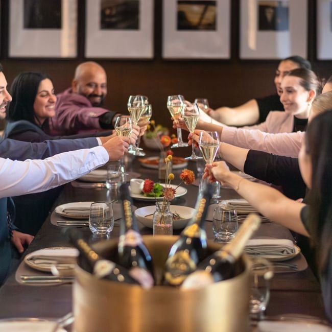 Group of friends toasting with champagne glasses at an elegant dinner table with framed artwork on the wall behind them