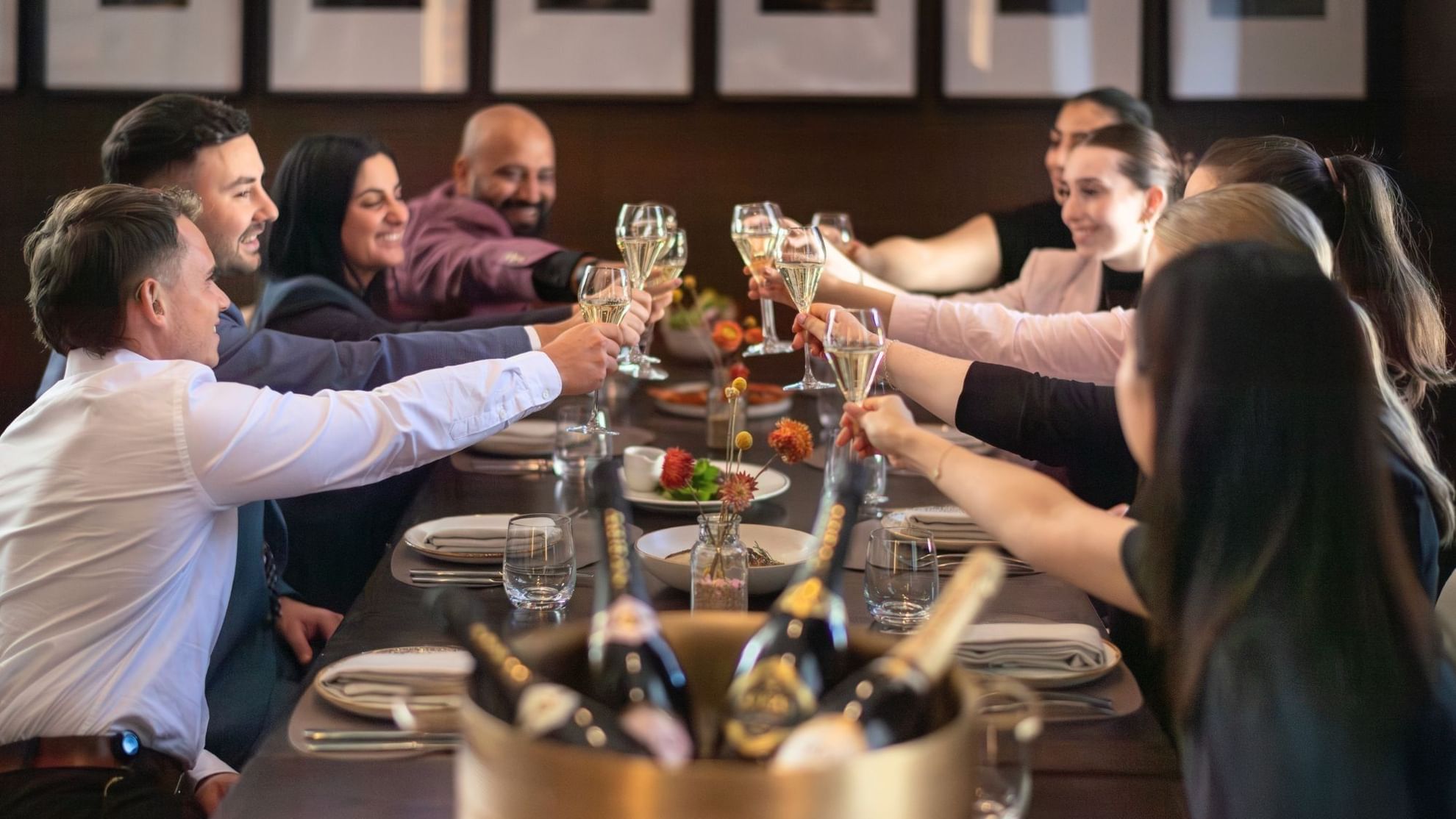 Group of friends toasting with champagne glasses at an elegant dinner table with framed artwork on the wall behind them