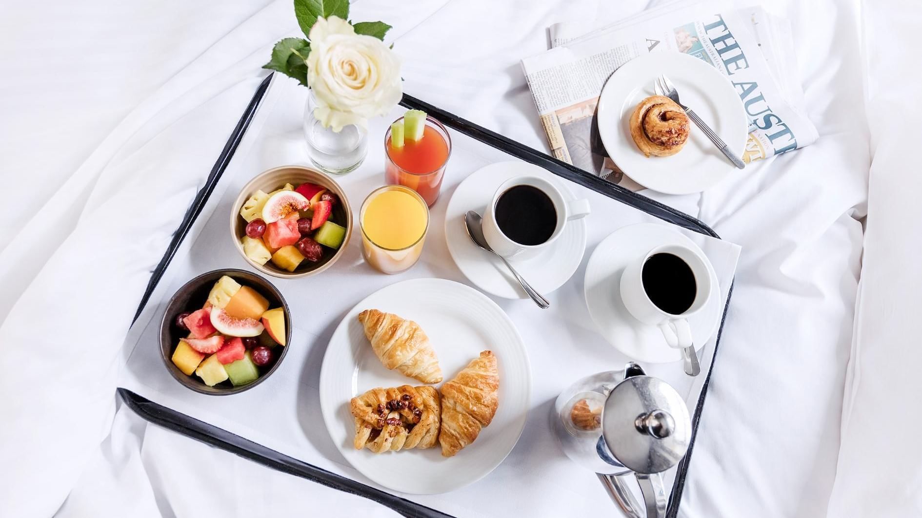 Paradox Sydney breakfast in bed with croissants, fruit salad, coffee, and newspaper on white bedspread.