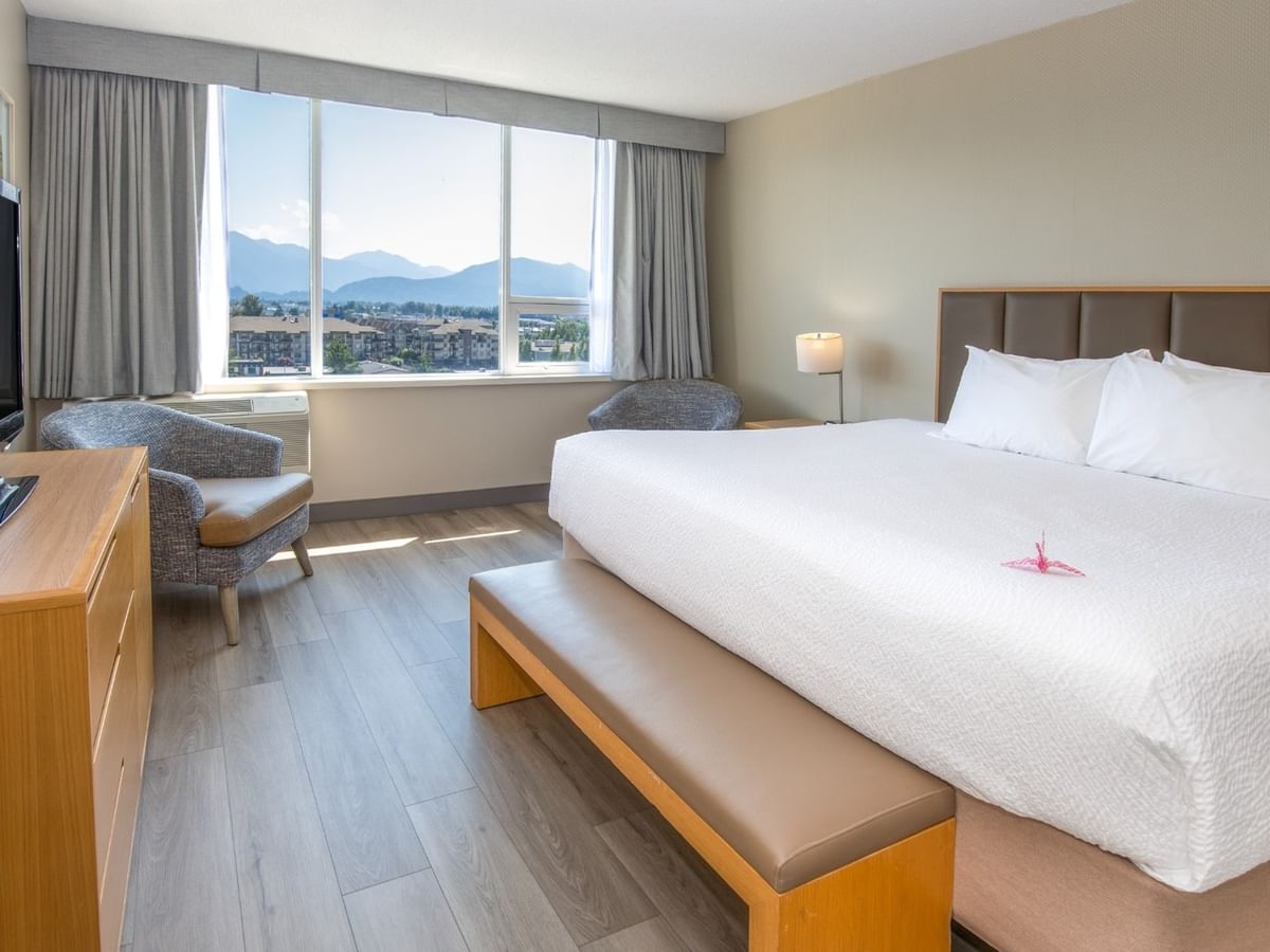 A hotel room with a large bed, chair, TV, dresser, and large window with mountain view.