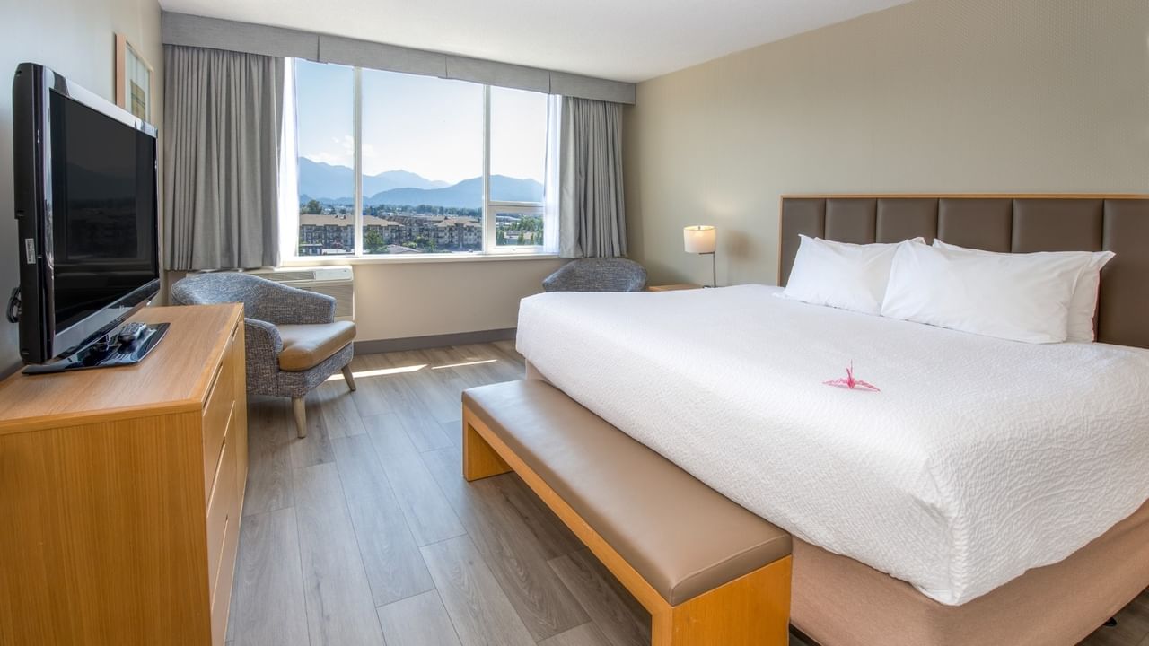 A hotel room with a large bed, chair, TV, dresser, and large window with mountain view.