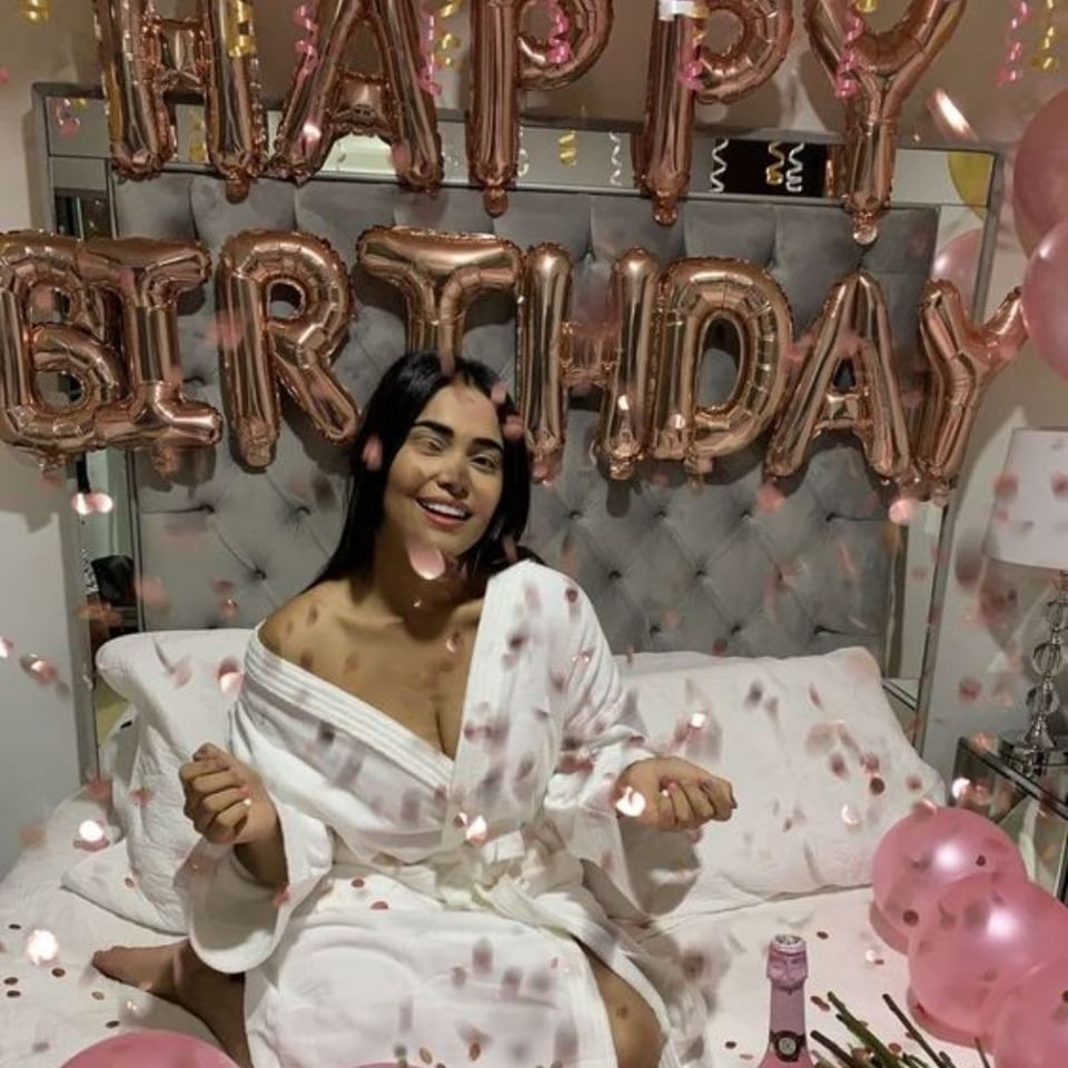Woman in white bathrobe sitting on a bed with rose gold Happy Birthday balloons and falling confetti at Hotel X Toronto