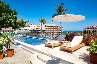 Two woven lounge chairs under a white umbrella sit next to an outdoor rooftop swimming pool at Casa Dona Susana