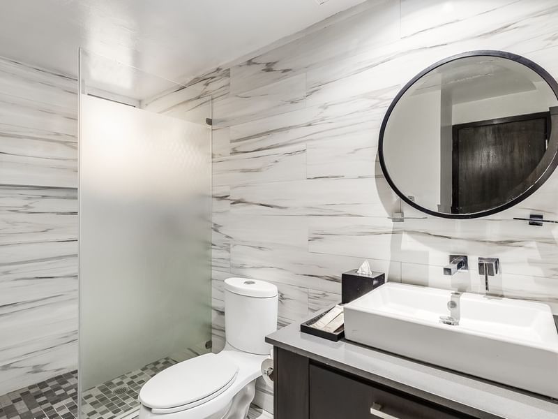 Deluxe room bathroom vanity with marble-patterned walls, a round mirror, and a frosted shower door at Sunvivia Mazatlán