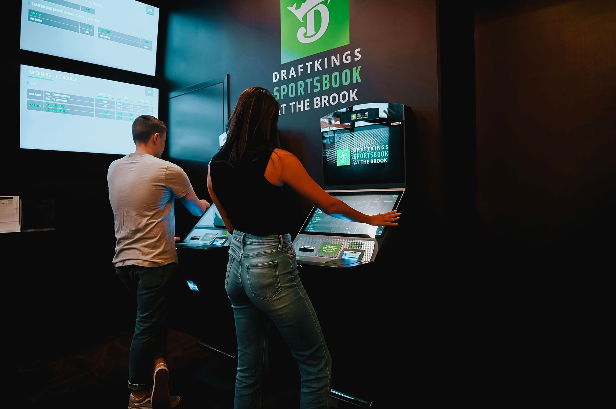 Guests use DraftKings Sportsbook kiosks beneath two digital sports betting screens inside the casino area at The Brook