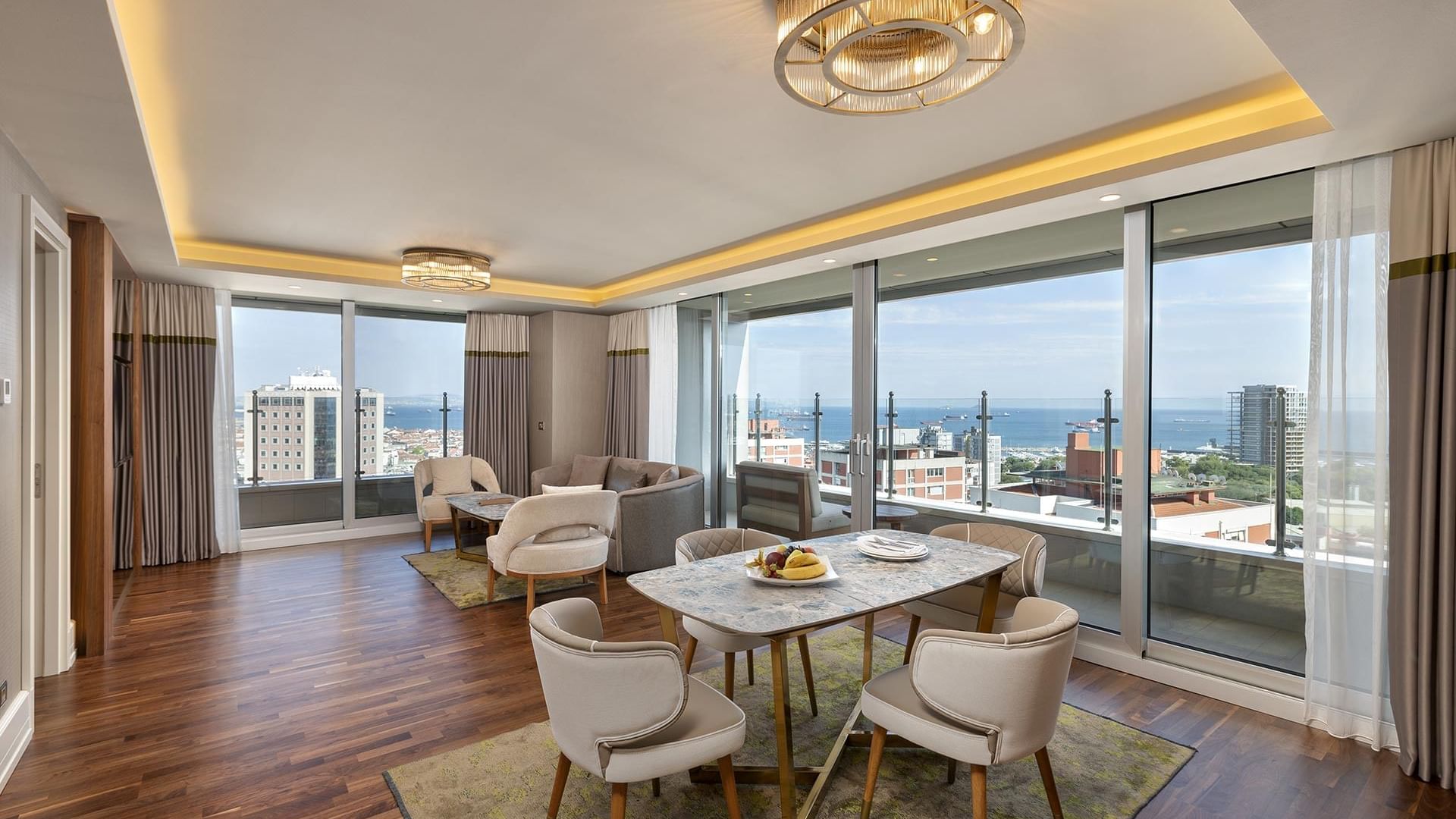 Titanic Port Bakırköy'de, şık mobilyalar ve dekoratif öğelerle donatılmış, yemek alanı, geniş pencereler ve şehir manzarası sunan Suite odada modern oturma alanı.
