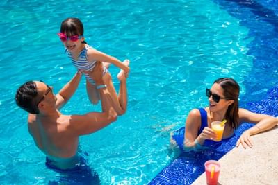 A family of three, including a young girl, enjoys swimming in a pool with drinks.
