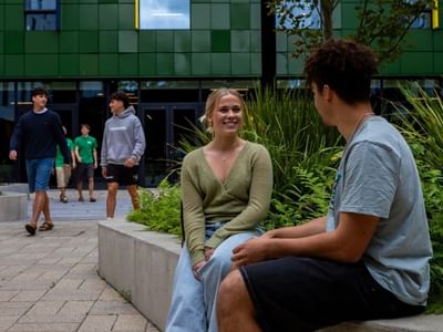 Two young people sitting on a concrete bench surrounded by greenery outside a modern building with green tiled facade.