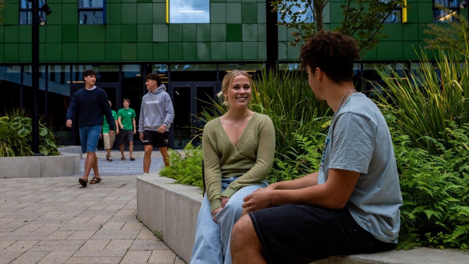 Two young people sitting on a concrete bench surrounded by greenery outside a modern building with green tiled facade.