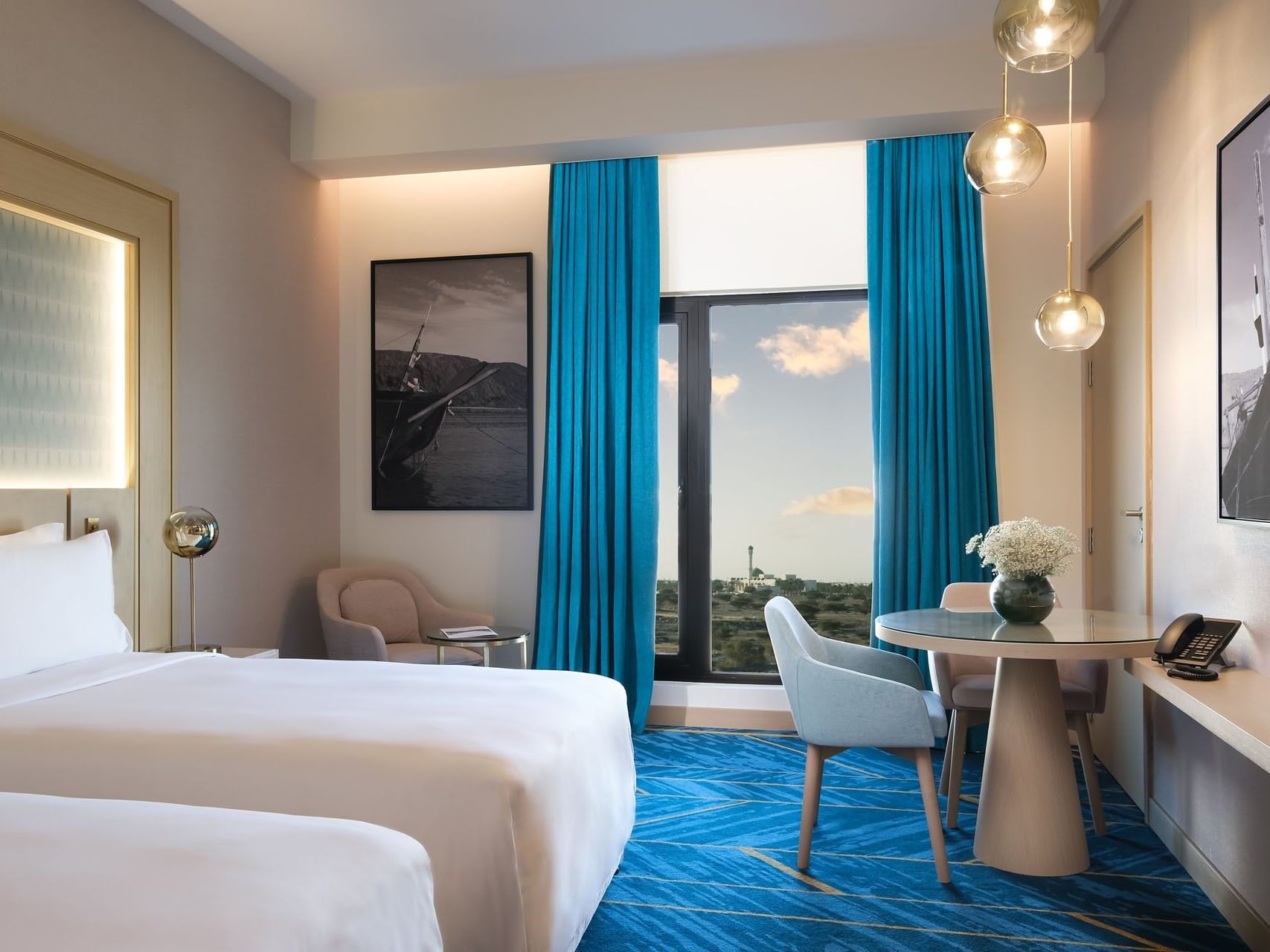 A hotel room with two beds, a dining table, and a window with blue curtains.