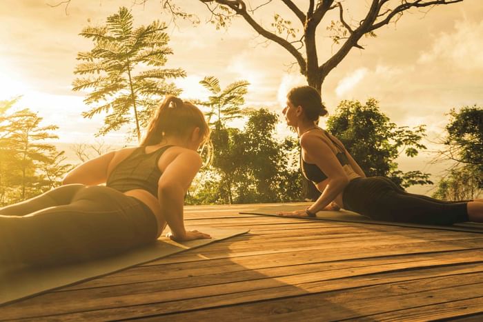 Two women practicing cobra yoga pose on mats over wooden outdoor deck during golden sunset hour at Los Altos Resort