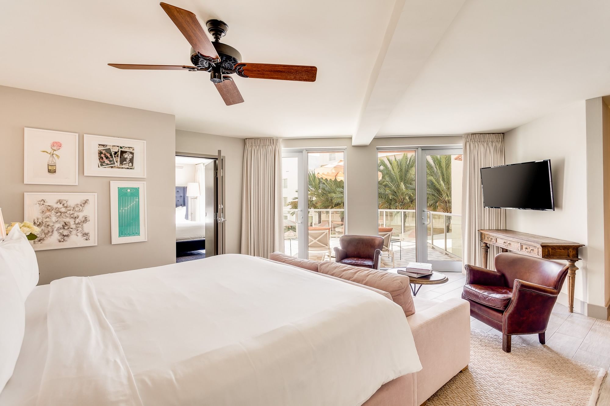 A wide view of a hotel room with a large bed, sofa, armchairs, and a balcony with palm trees.