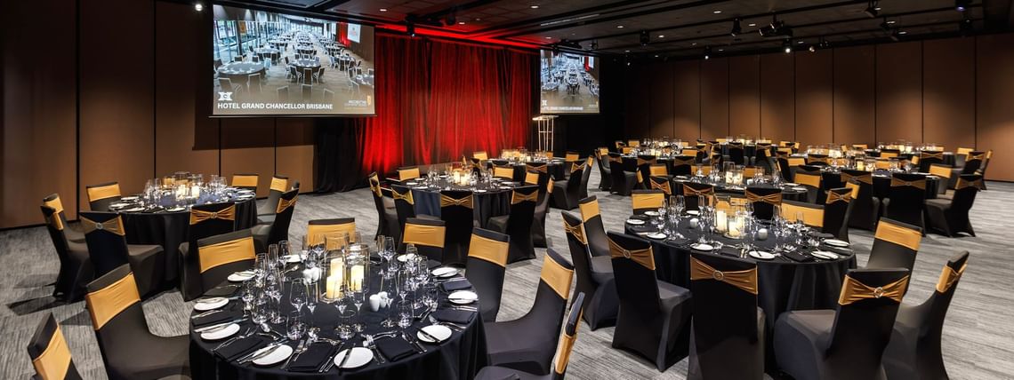 Large corporate gala dinner setup with round banquet tables, stage lighting, and presentation screens at Hotel Grand Chancellor Brisbane event venue.