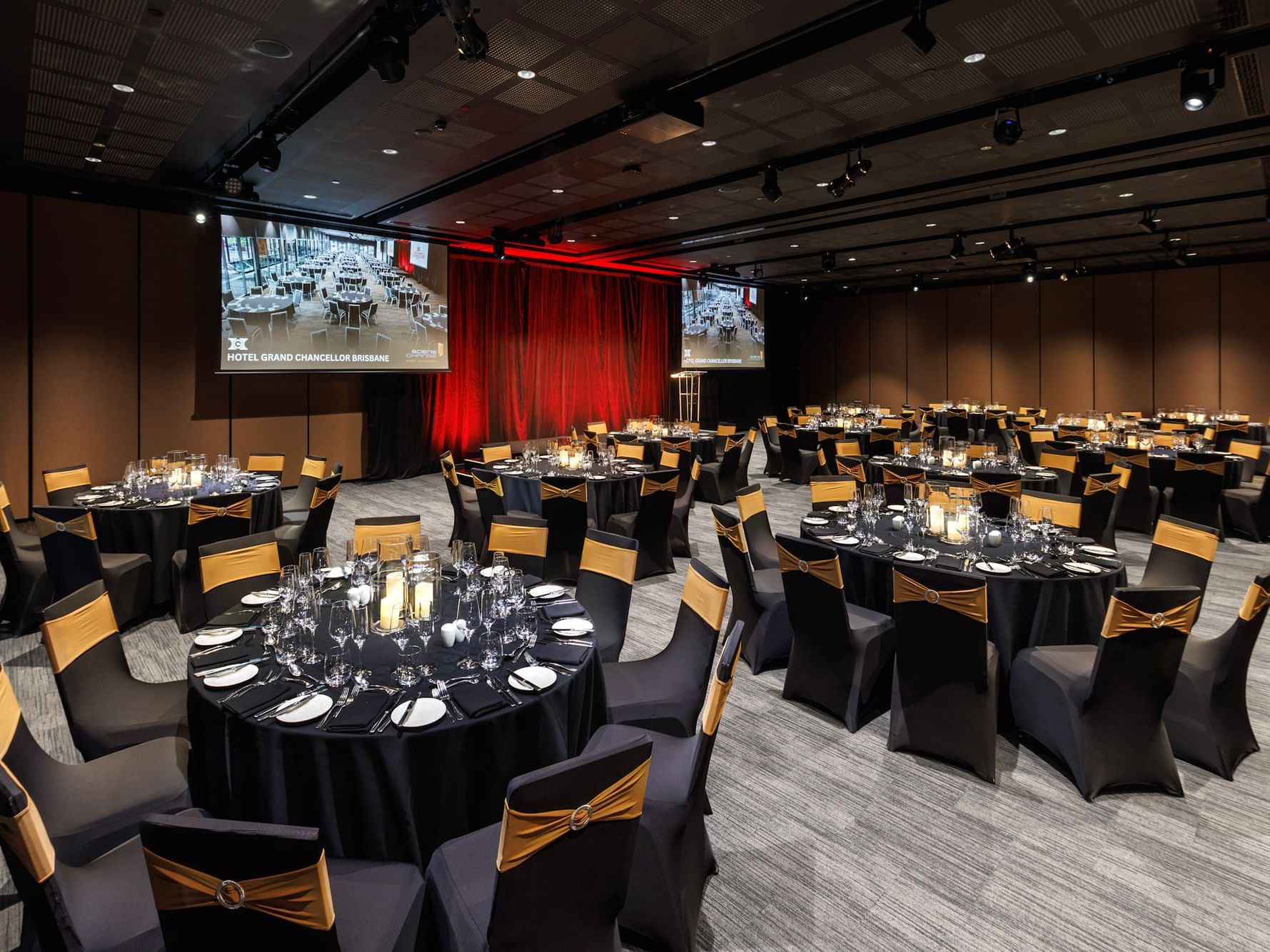 Large corporate gala dinner setup with round banquet tables, stage lighting, and presentation screens at Hotel Grand Chancellor Brisbane event venue.