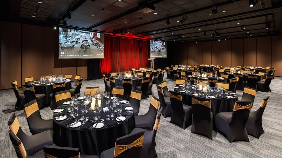 Large corporate gala dinner setup with round banquet tables, stage lighting, and presentation screens at Hotel Grand Chancellor Brisbane event venue.