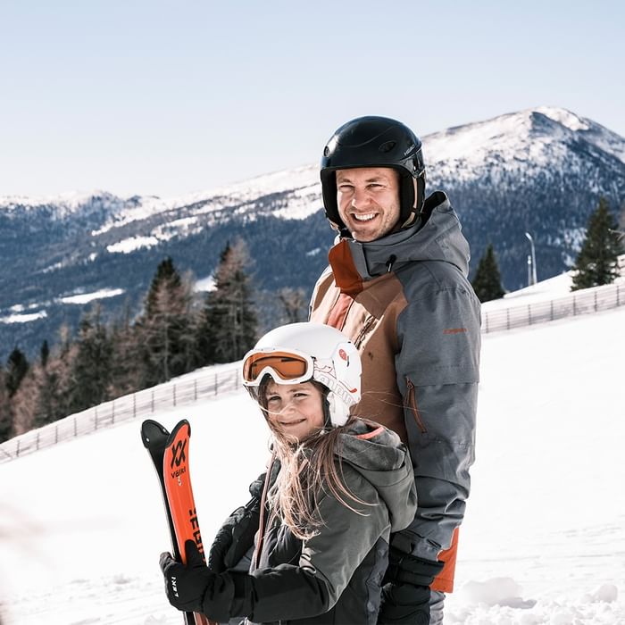 Father and daughter in winter gear smiling on a snowy mountain slope with skis.