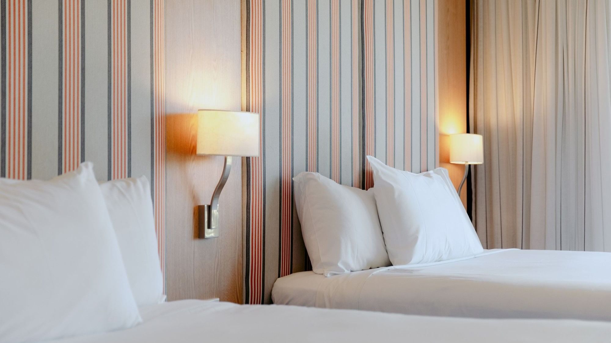 Two beds with white sheets and pillows, flanked by lit wall lamps against striped wallpaper.