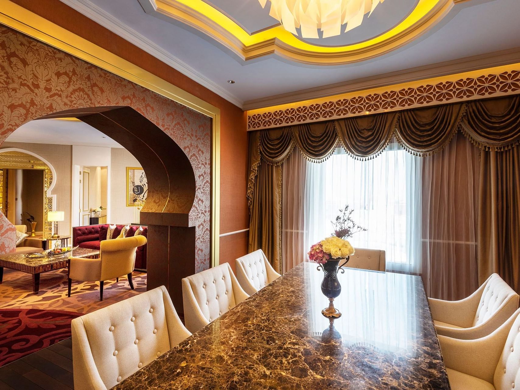 Elegant dining room with marble table, white chairs, and floral centerpiece, adjacent to a lounge area with ornate decor.
