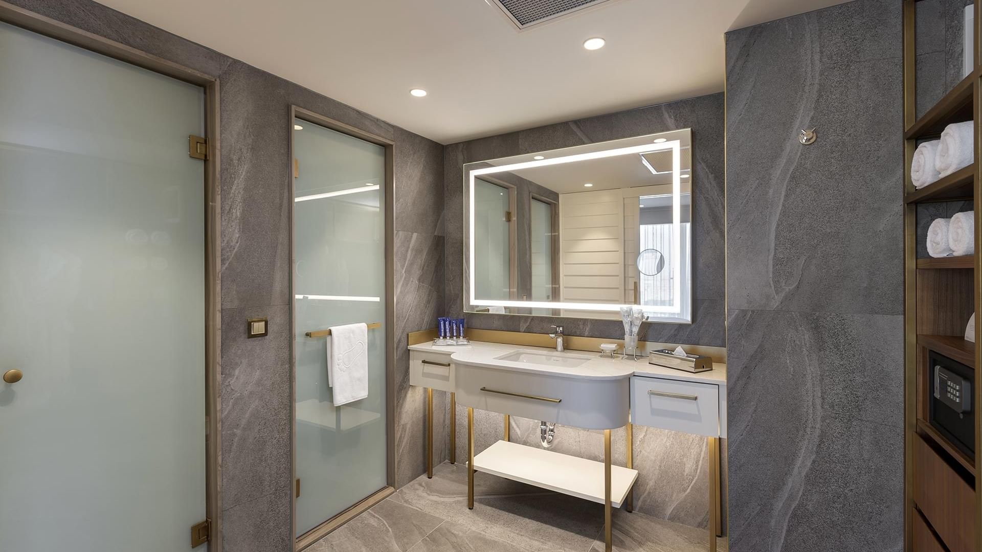 Titanic Port Bakırköy'de, büyük aynalı, lavabolu, cam duş kapılı, fayanslı duvarlı ve havlularla dolu ahşap raflı Suite odada modern banyo.