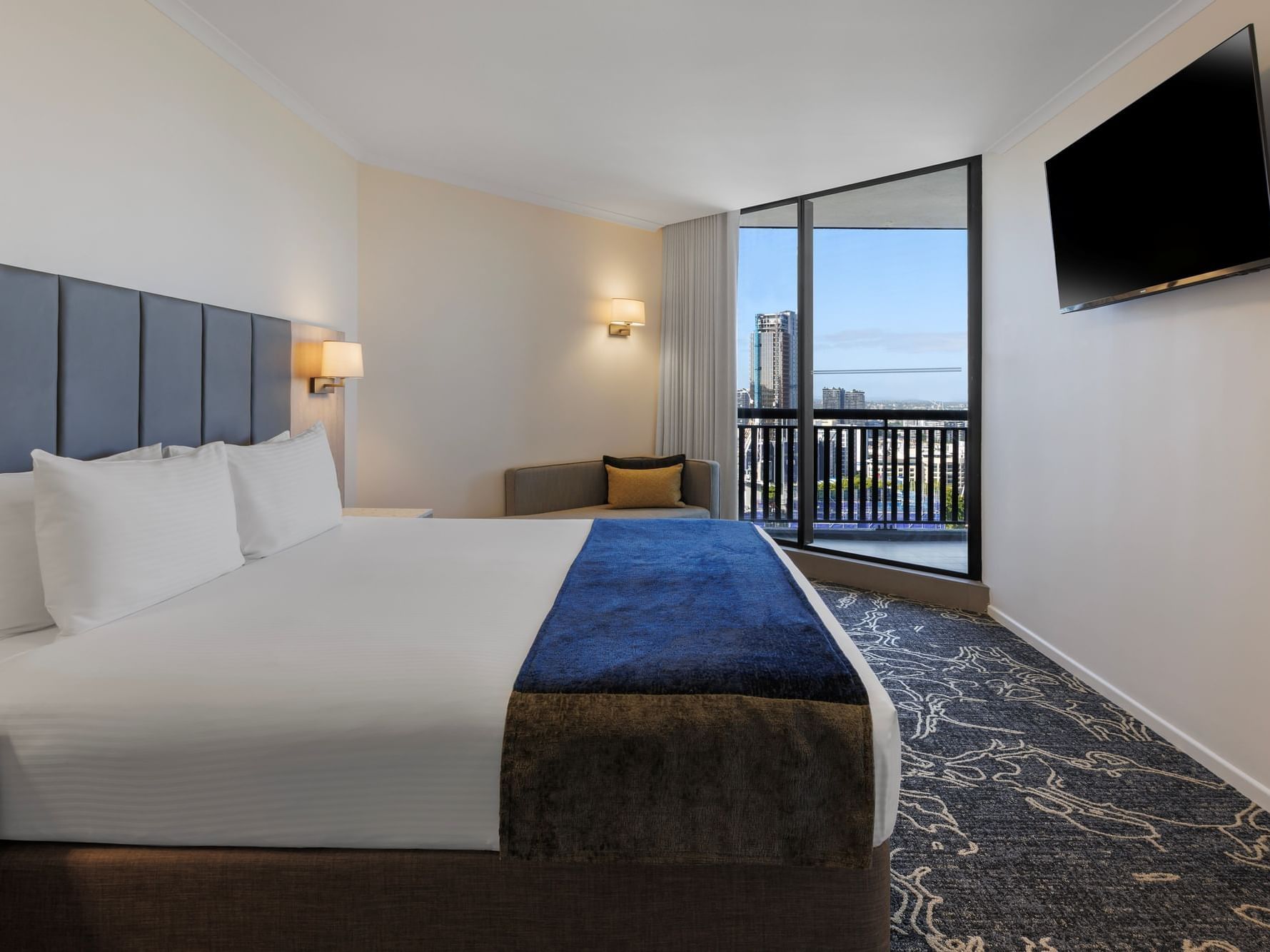Modern Superior King Suite Bedroom at Hotel Grand Chancellor Brisbane featuring a king bed with a blue velvet runner, a private balcony with city views, and a wall-mounted flat-screen TV.
