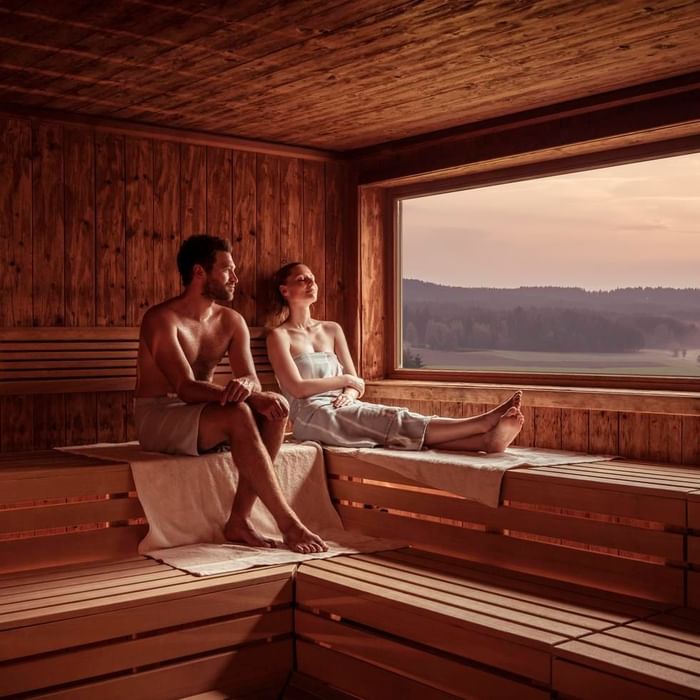 Paar entspannt in einer hölzernen Sauna mit Blick auf eine ländliche Landschaft bei Sonnenuntergang.