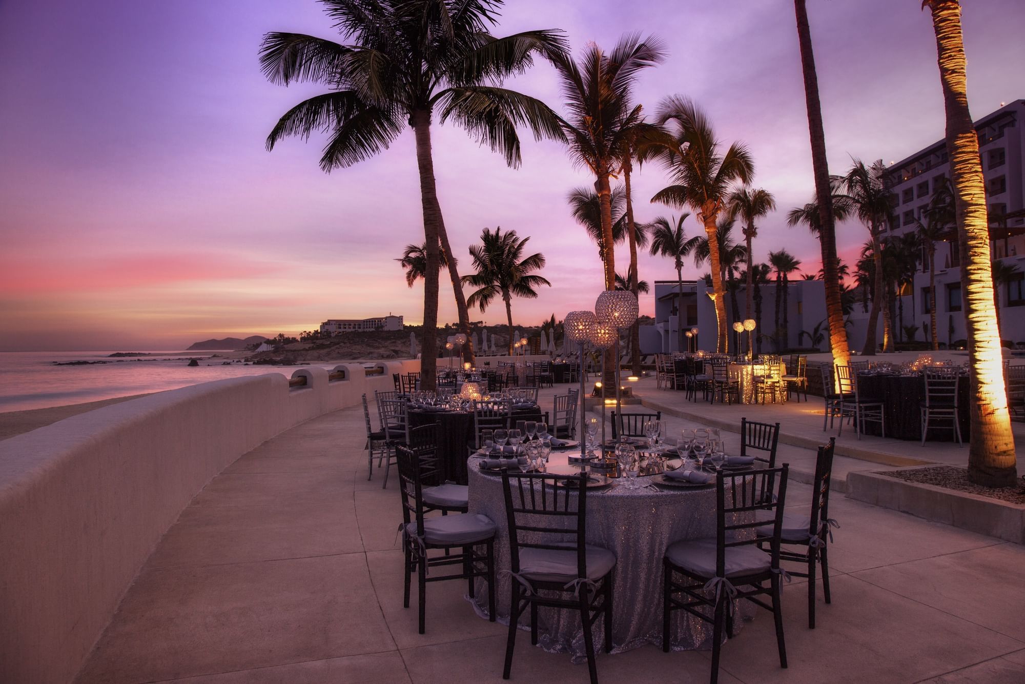 Outdoor dining setup at twilight, with palm trees and a stunning coastline at Marquis Los Cabos