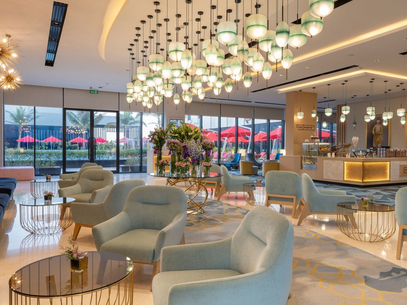 A luxurious lobby with blue chairs, glass tables, floral arrangements, and a large chandelier with green shades.
