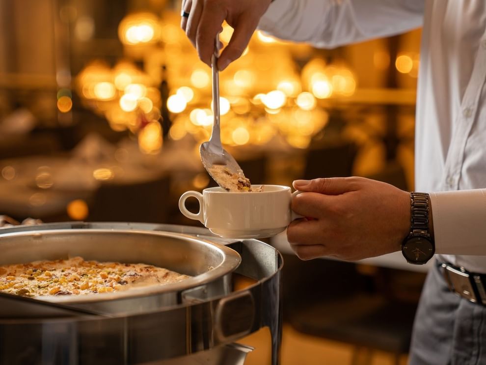 Person ladles dessert topped with nuts into white cup in Al Firdous Restaurant at Saja Warwick Madinah