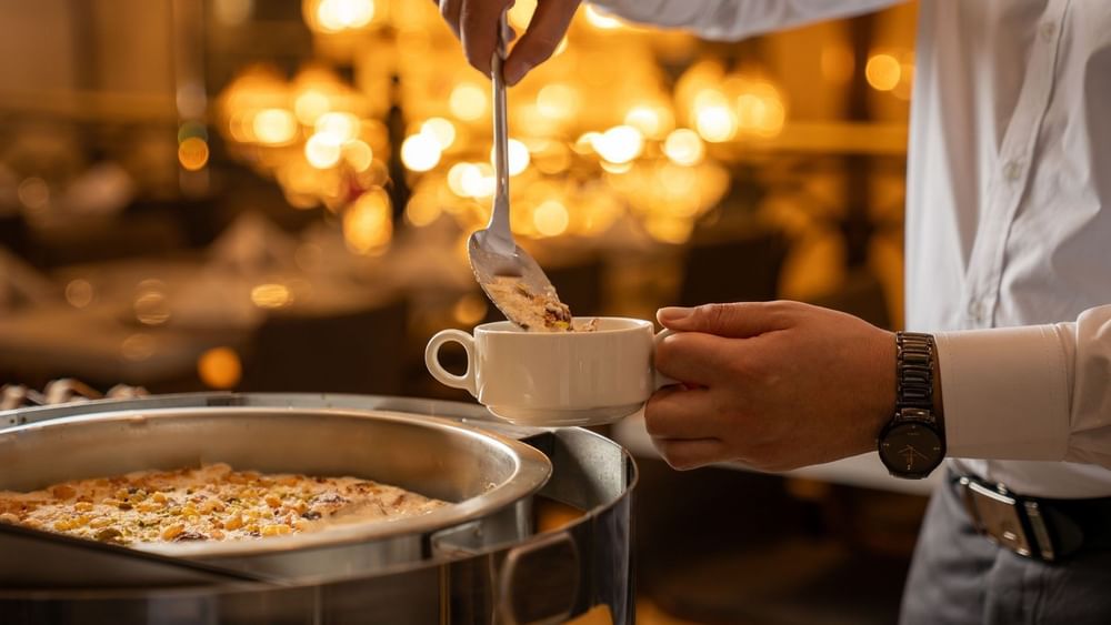 Person ladles dessert topped with nuts into white cup in Al Firdous Restaurant at Saja Warwick Madinah