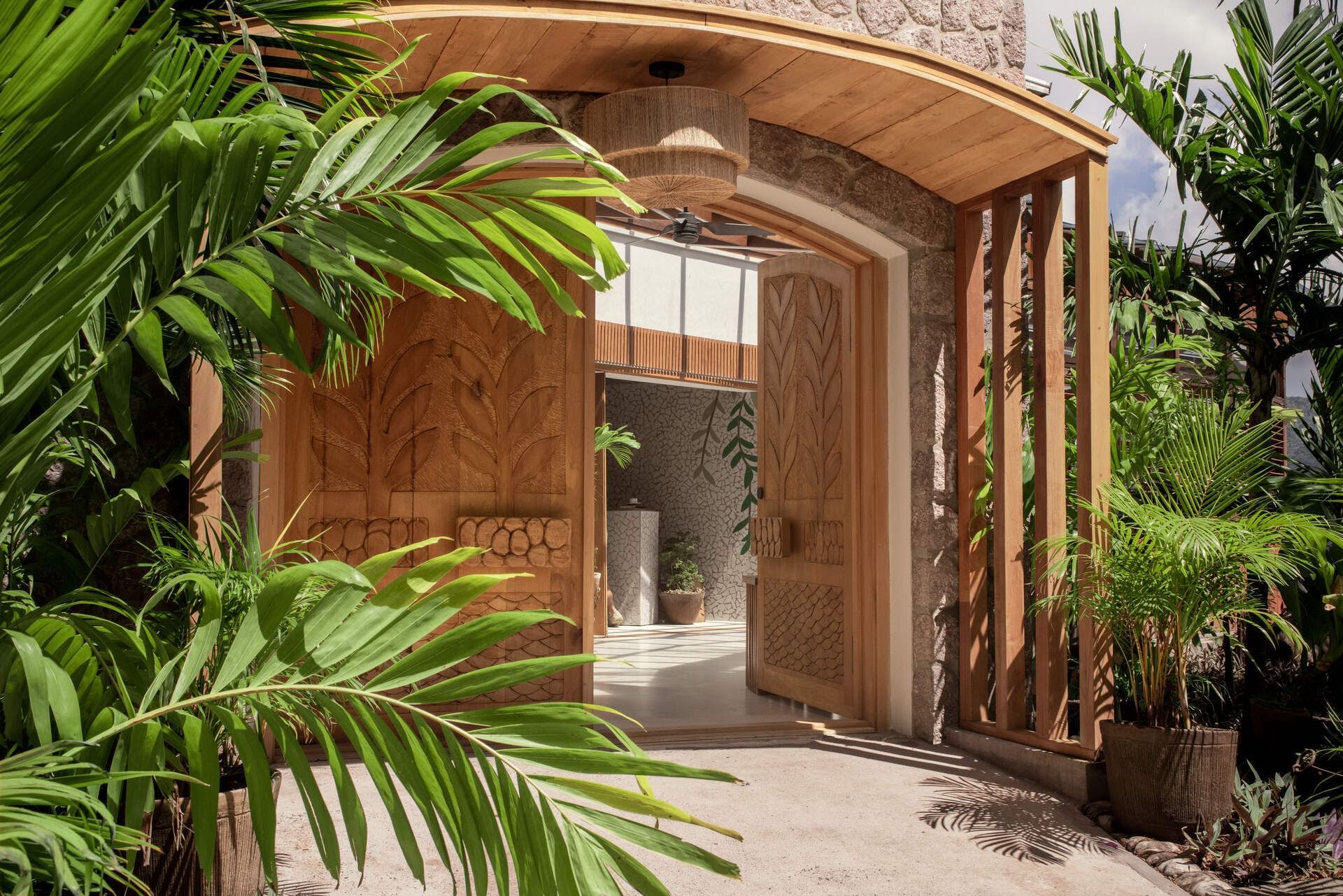 Wooden entrance archway with carved doors and tropical plants on both sides, leading to an indoor space.