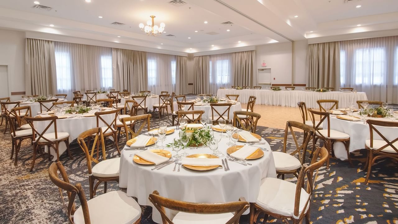 Banquet hall with round tables, white cloths, wooden chairs, and floral centerpieces under a chandelier.