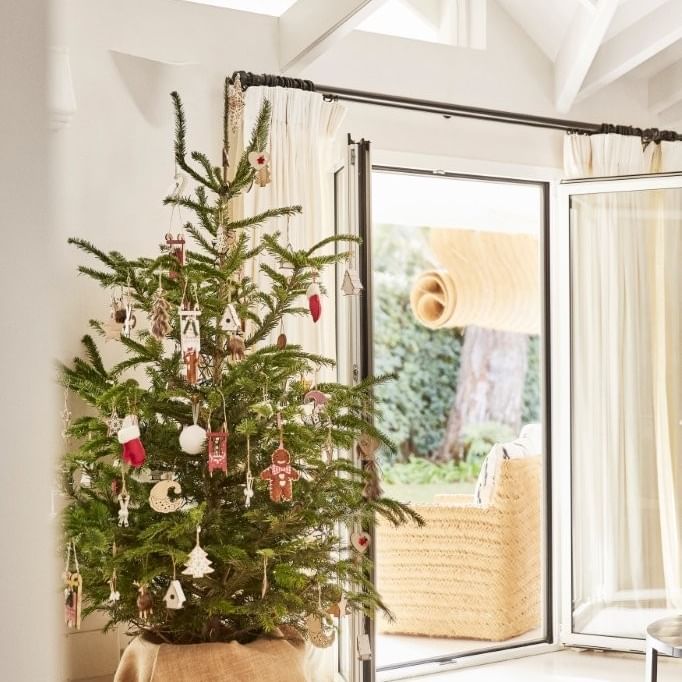 Decorated Christmas tree in a burlap sack, in a bright room with white walls and a glass door.