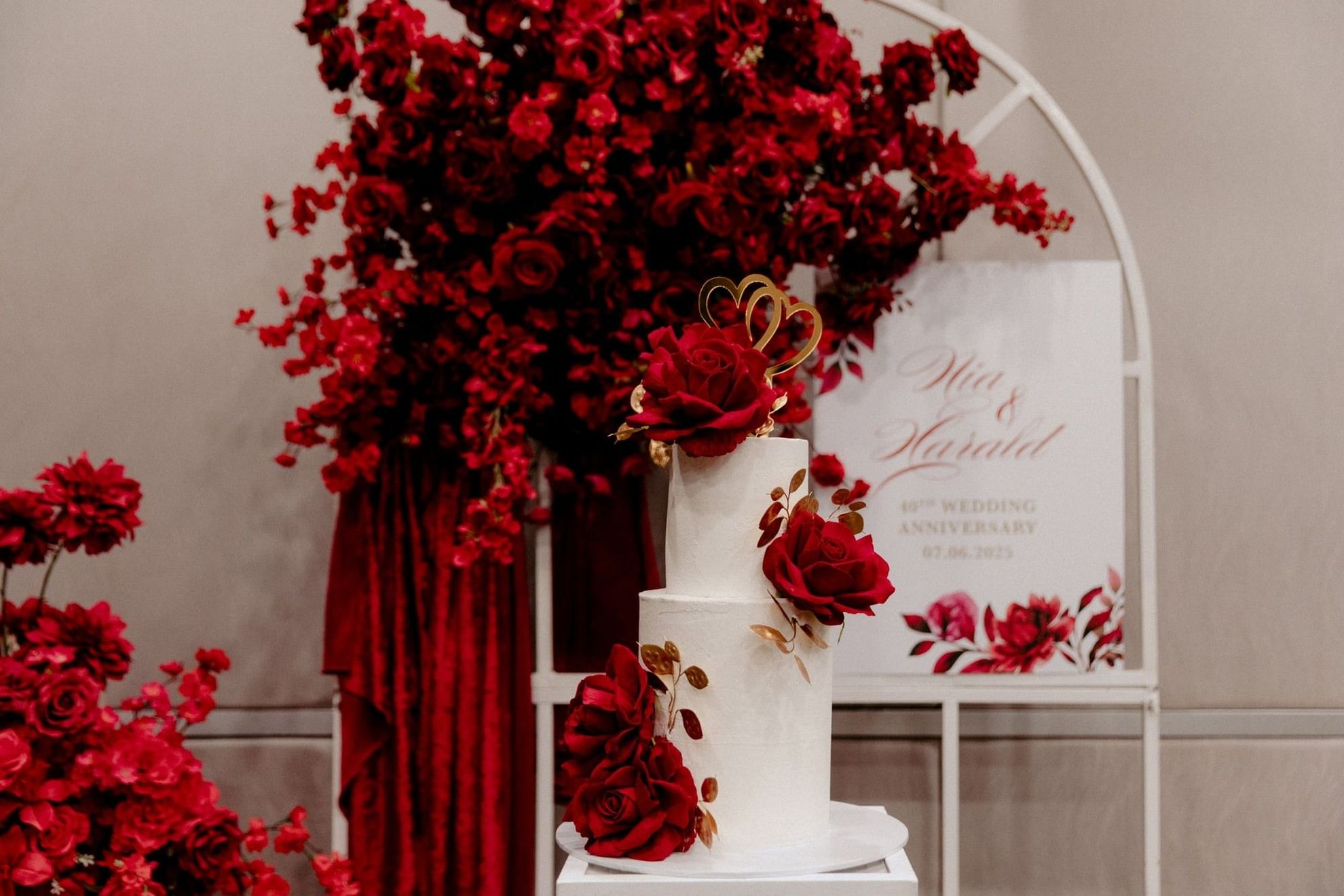 White wedding cake with red roses and gold accents surrounded by red flowers