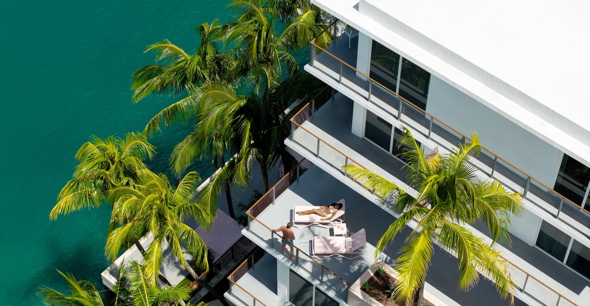 High-angle view of guest lounging on a private balcony with palm trees facing water at Grand Beach Hotel Bay Harbor