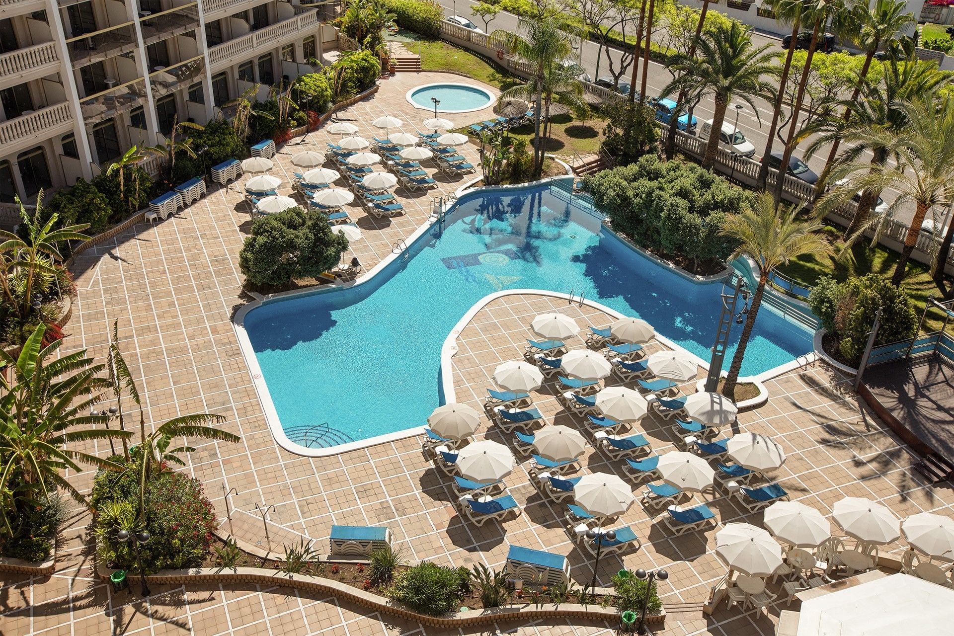 Aerial view of a tropical resort's blue lagoon-style pool, surrounded by many white umbrellas and lush green palm trees.