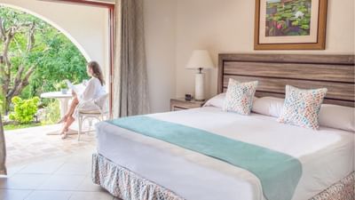 Woman sitting on a terrace with a plush king bed and a wall painting visible inside at Sugar Cane Club Hotel & Spa