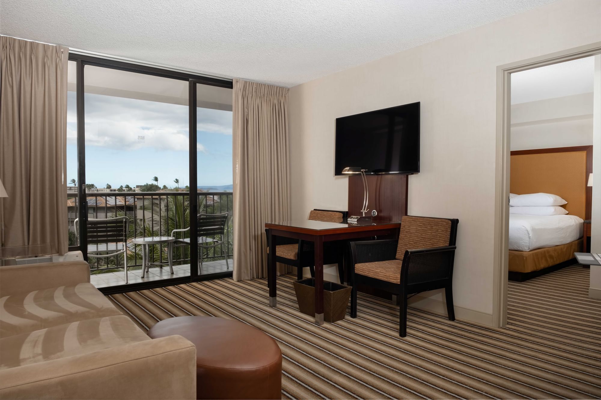 A hotel room with a desk, chair, TV, sofa, and balcony with a view.