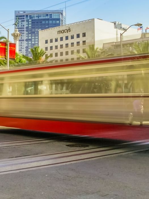 Motion-blurred streetcar passes palm trees and urban buildings near Macys at Union Square.